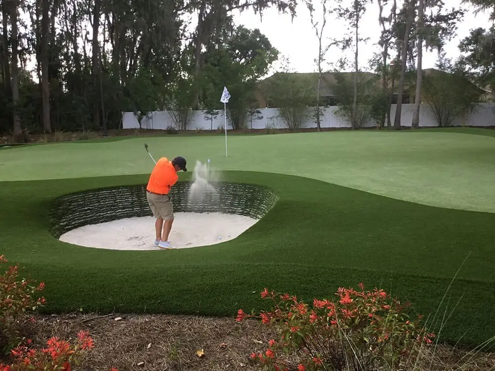 Signature backyard putting green with synthetic turf, stacked-sod bunkers, and wooded landscaping by Celebrity Greens®