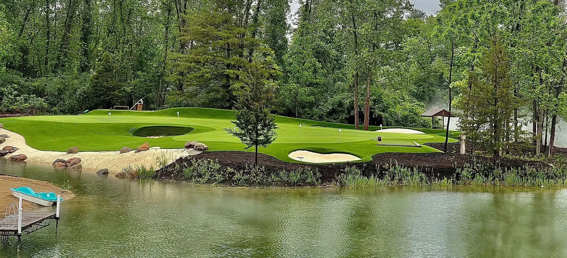 Signature lakeside putting green with synthetic turf, sculpted bunkers, and wooded backdrop by Celebrity Greens®